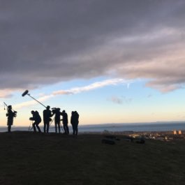 A silhouetted production crew using Zoom equipment to capture audio atop a hill overlooking a coastal village.